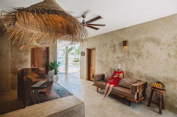 A modern living room with a laptop on a wooden table, a sofa, a ceiling fan, and a view of a pool. - Passion Boutique Hotel Zanzibar (Jambiani)