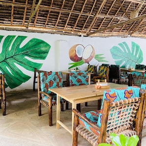 A restaurant with a thatched roof, wooden tables, and chairs with colorful fabric. - Max Hotel Nungwi (Nungwi)
