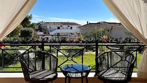 Un balcon avec deux fauteuils en osier et une petite table, donnant sur un jardin et des immeubles.
