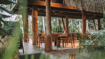 An outdoor dining area with wooden tables and chairs, surrounded by large trees and a thatched roof structure.