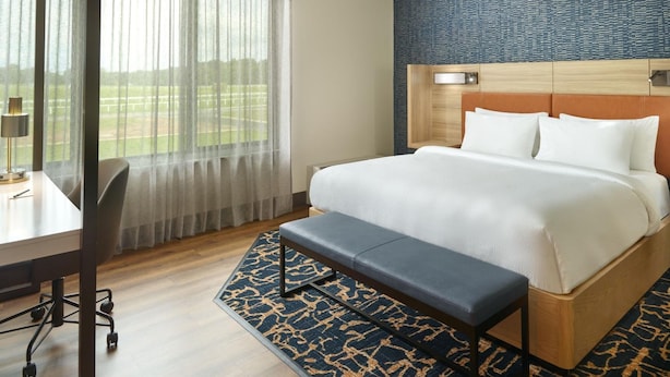 Quarto de hotel com uma cama grande, uma escrivaninha com cadeira, um banco e vista para um campo de golfe através de cortinas finas.