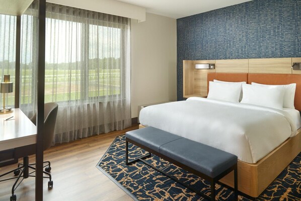 A hotel room with a large bed, a desk with a chair, a bench, and a view of a golf course through sheer curtains. - Springhill Suites Franklin Mint (South Portland)