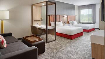 A hotel room with a desk, two beds, a sofa, and a large window.