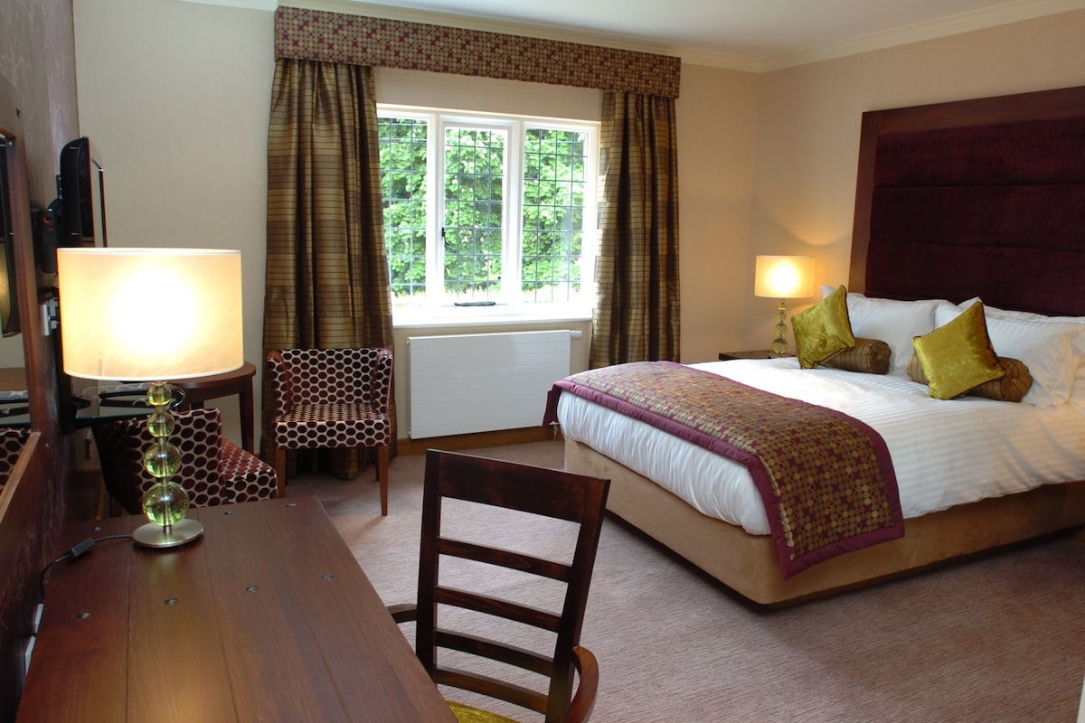 A hotel room with a bed, two bedside lamps, a chair, a desk, and a window with curtains.