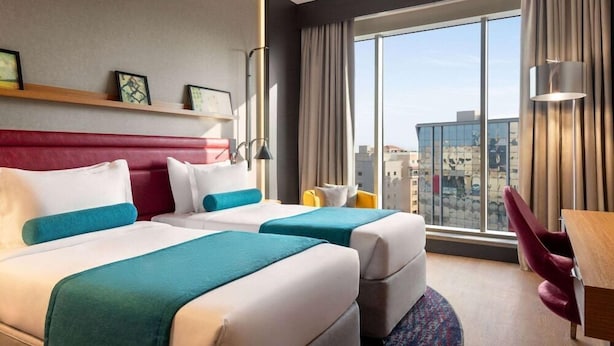 A hotel room with two beds, a desk, a chair, a large window with a city view, and a red headboard.