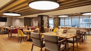 A modern dining area with multiple tables and chairs, large circular ceiling lights, and large windows offering a view of the cityscape.