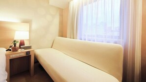 Een hotelkamer met een bank, een nachtkastje met een lamp en een raam met dunne gordijnen.