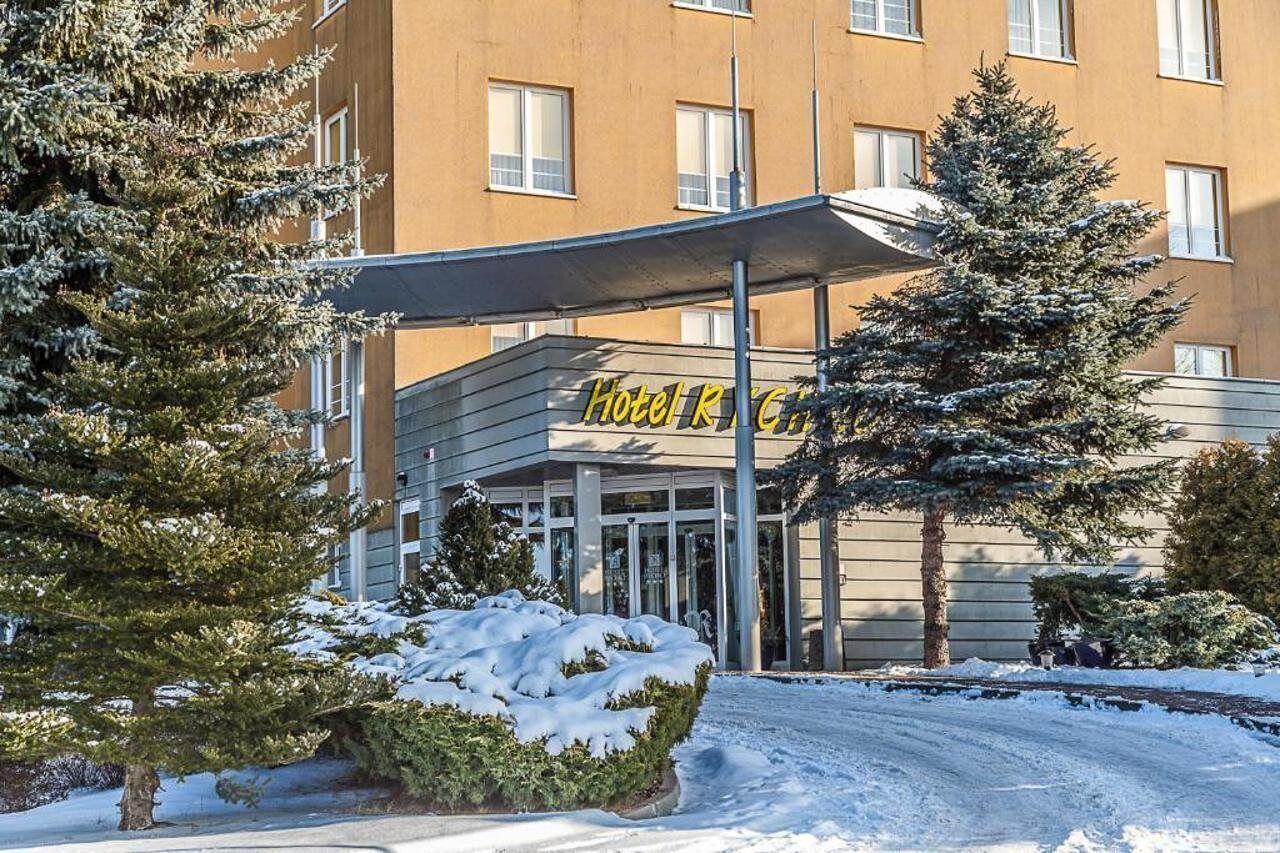 Entrada de hotel com marquise, árvores cobertas de neve e céu claro.