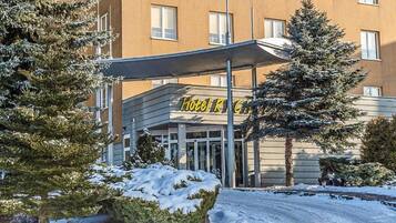 Entrada de hotel com marquise, árvores cobertas de neve e céu claro.