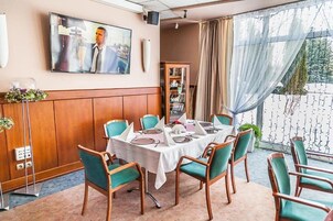 Un comedor con una mesa redonda dispuesta para seis personas, sillas teal alrededor, un televisor montado en la pared y vistas a un balcón con cortina.