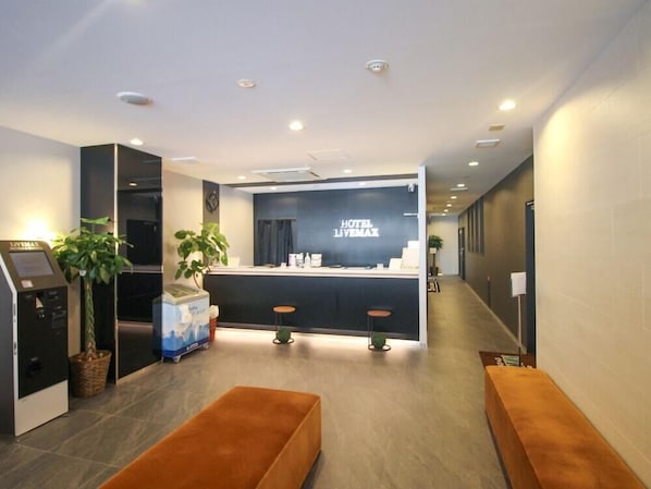 Uma área de recepção de hotel com design moderno, incluindo um balcão de atendimento, espaço de estar e um quiosque de autoatendimento.