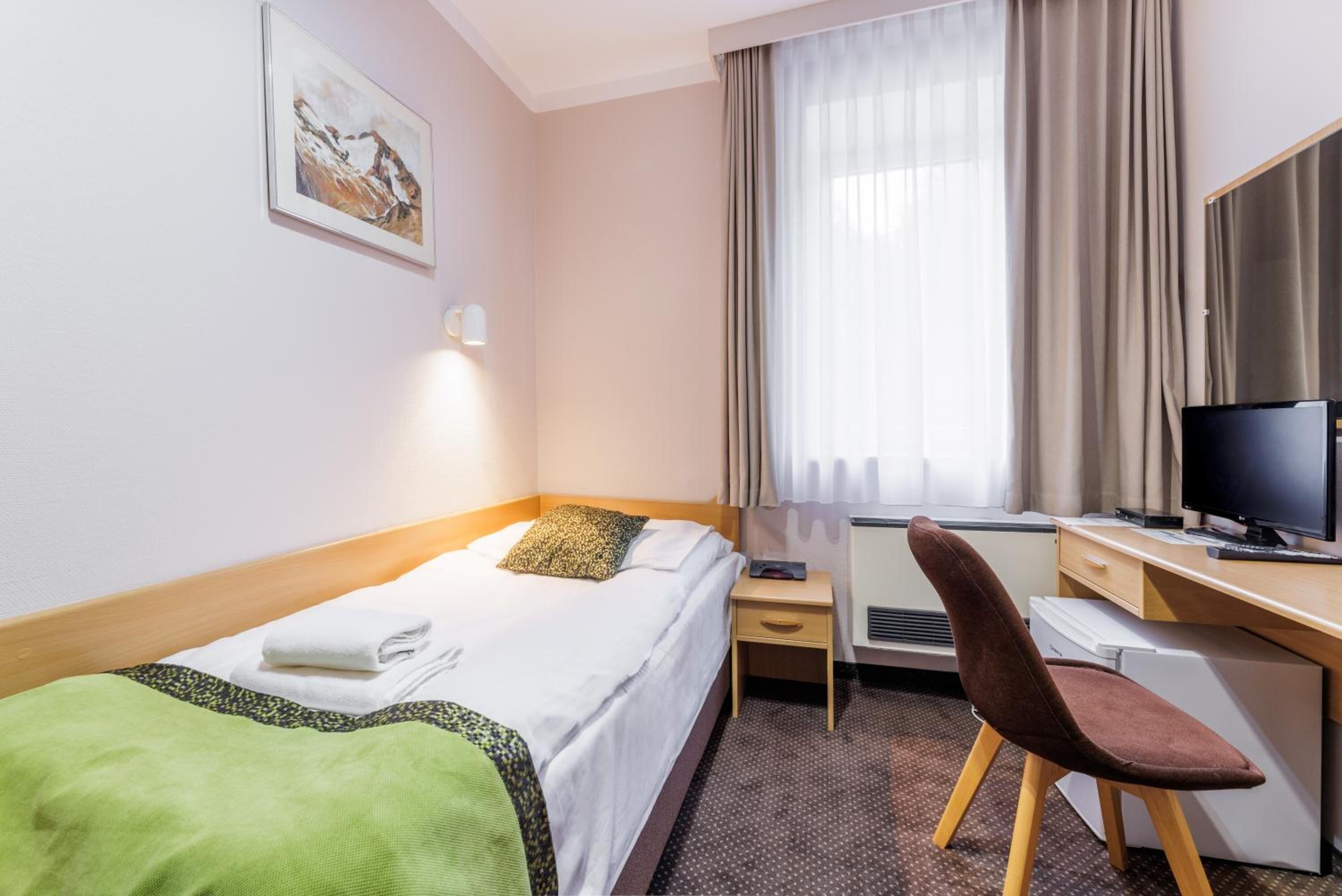A hotel room with a bed, a desk with a computer, a chair, a window with curtains, and a painting on the wall.