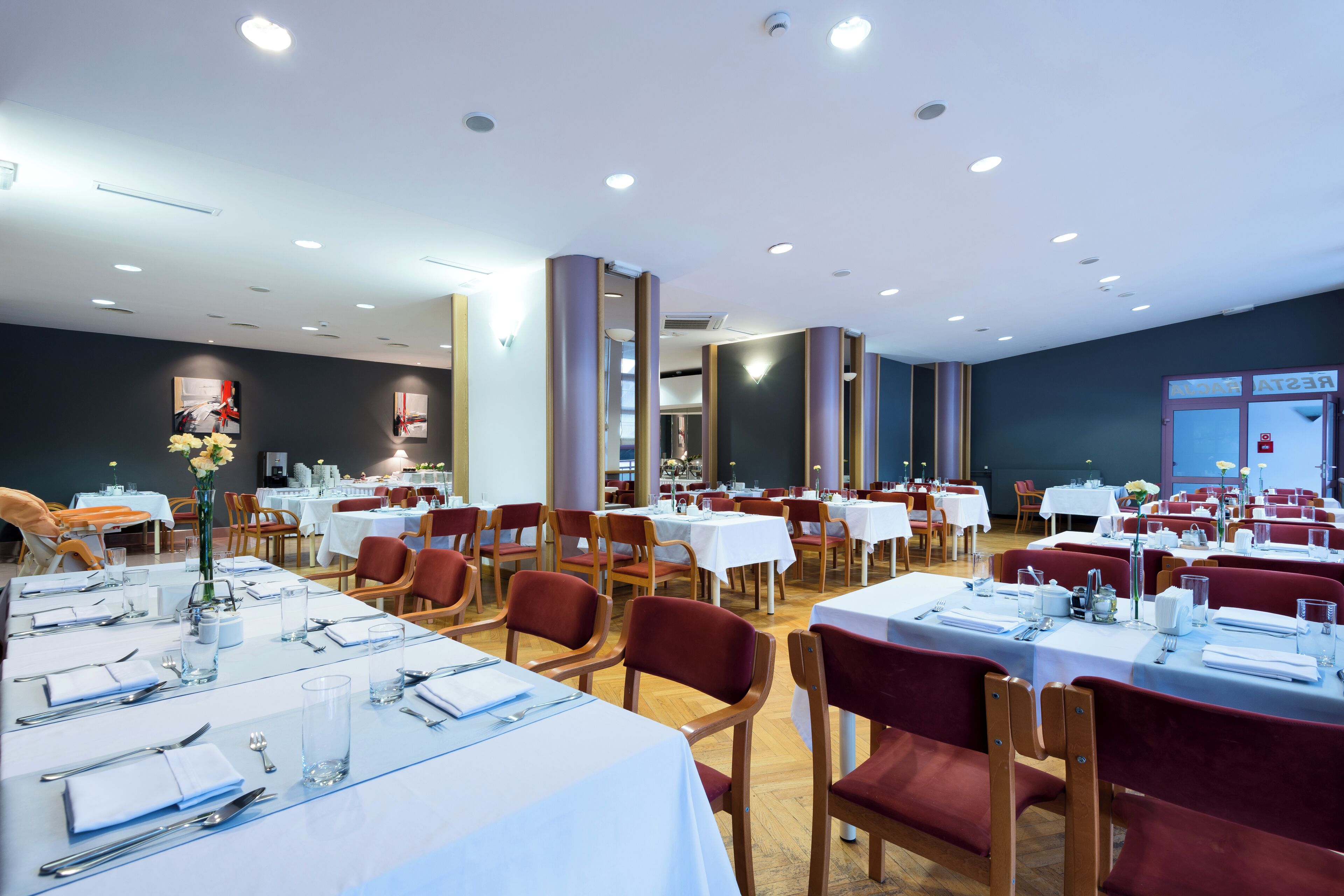 A spacious dining area with multiple tables and chairs, arranged in a modern setting with a high ceiling and recessed lighting.