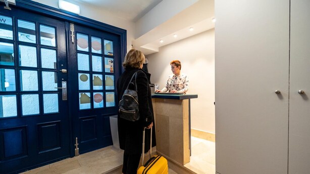Zona de recepción de un hotel con una puerta azul, un mostrador de recepción y una persona al lado de una maleta amarilla.
