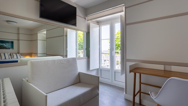 Ein modernes Wohnzimmer mit einer Couch, einem Holztisch, einem an der Wand montierten Fernseher und einem großen Fenster mit Blick auf Grün.