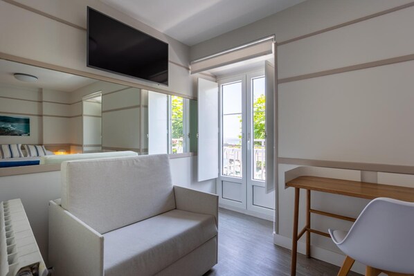 A modern living room with a sofa, a wooden table, a TV mounted on the wall, and a large window with a view of greenery outside. - Hotel El Puerto (Mundaka)
