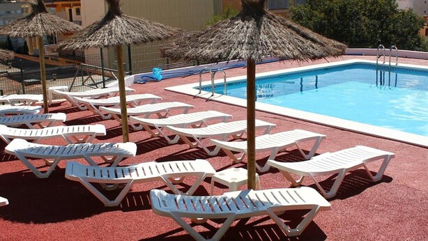 Une piscine entourée de bâtiments, avec des chaises longues et des parasols en chaume.