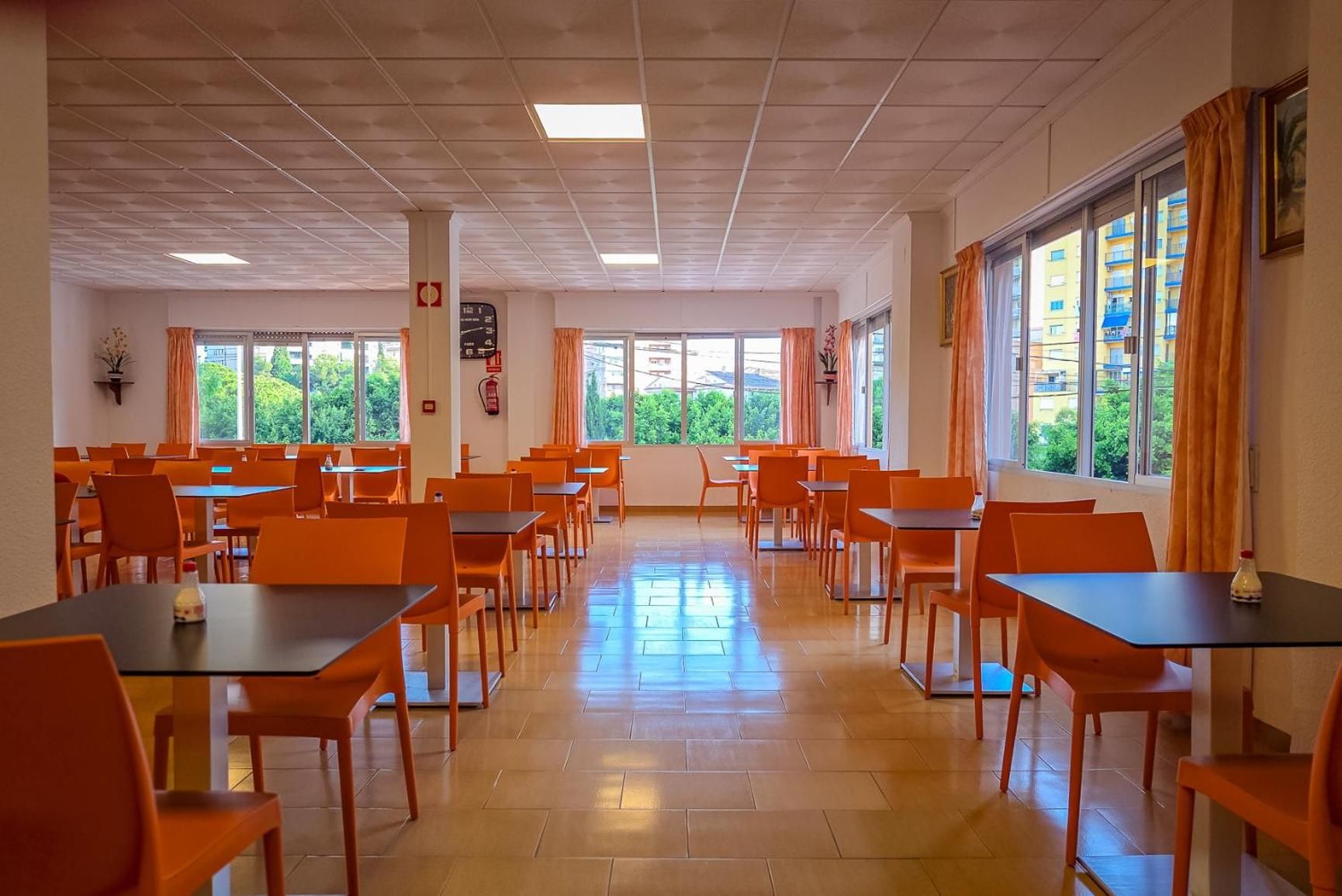 Un comedor espacioso con múltiples mesas y sillas, ventanas amplias con cortinas, y piso de baldosas.