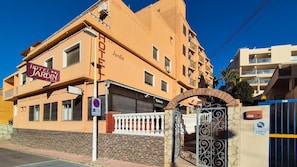 Hotel 'Hotel Jardin' con entrada protegida por una reja, señal de 'no estacionamiento' y un café a la vista.