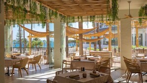 An outdoor dining area with tables and chairs, surrounded by large windows offering views of palm trees and the ocean. - Daia Slow Beach Hotel Conil - Adults Only (Conil de la Frontera)