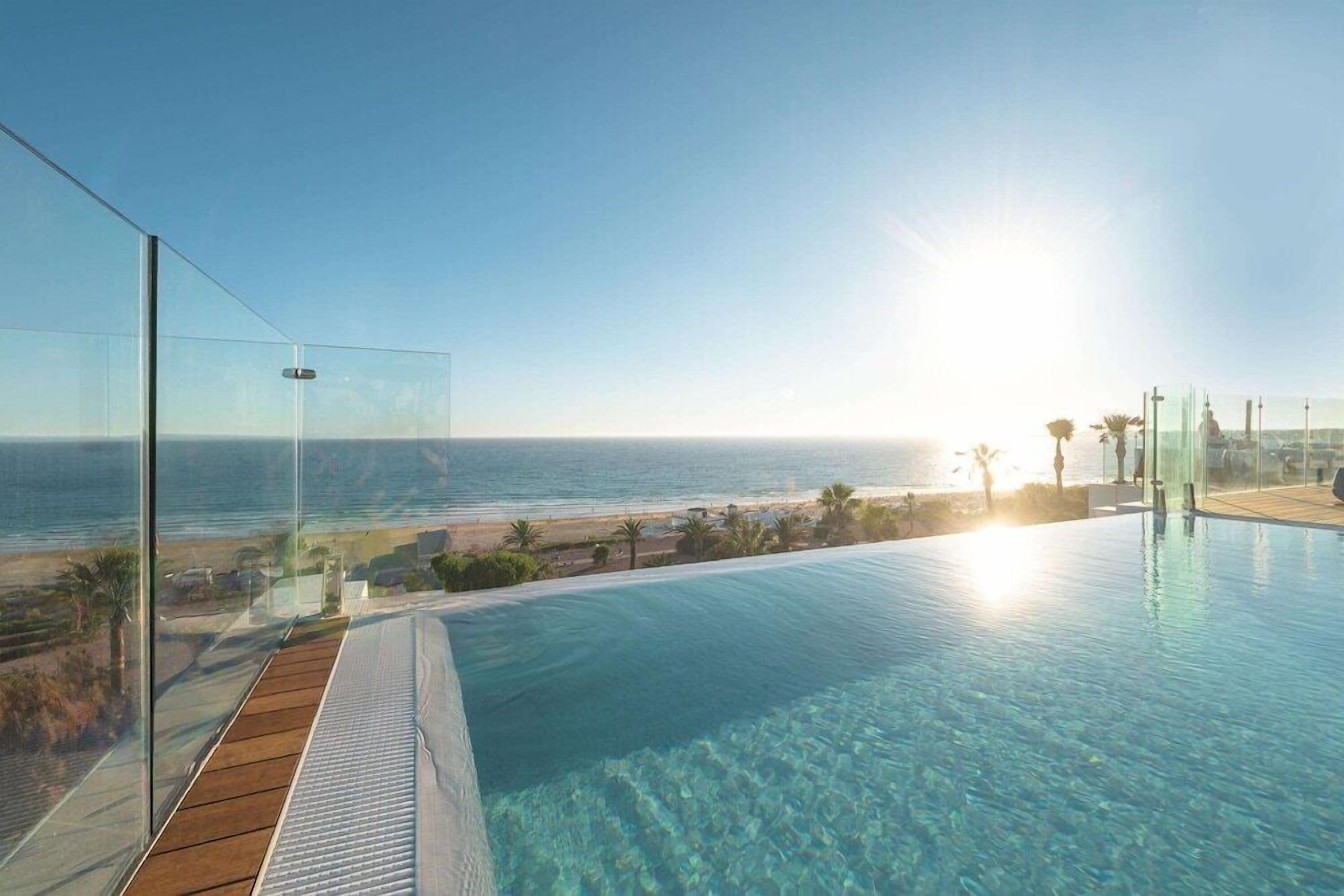 A clear view of a swimming pool with a wooden deck, overlooking a beach and the ocean.