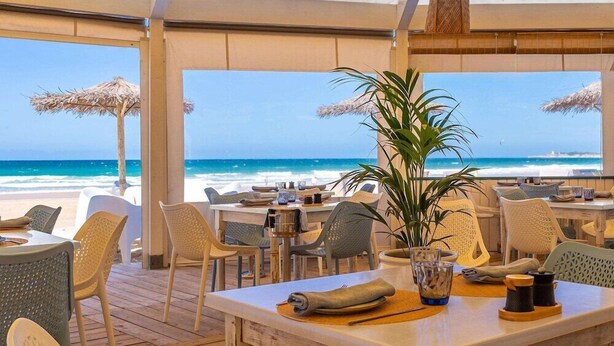 An outdoor dining area with tables and chairs, a large potted plant, and a view of the ocean. - Daia Slow Beach Hotel Conil - Adults Only (Conil de la Frontera)
