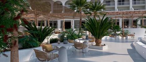 A spacious outdoor area with multiple seating arrangements, palm trees, and a building with arched windows in the background.