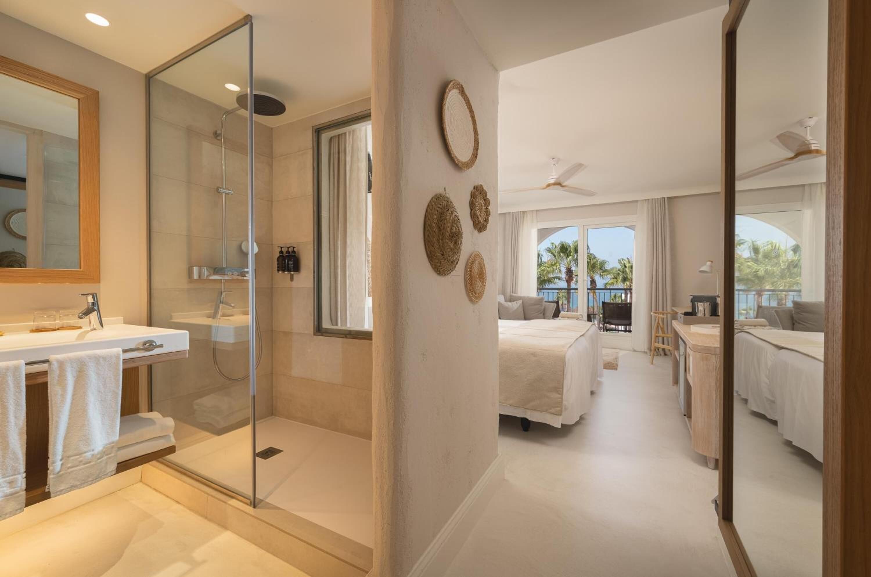 Une chambre d’hôtel moderne dotée d’une douche en verre, d’un grand lit, d’un bureau et offrant une vue sur les palmiers.