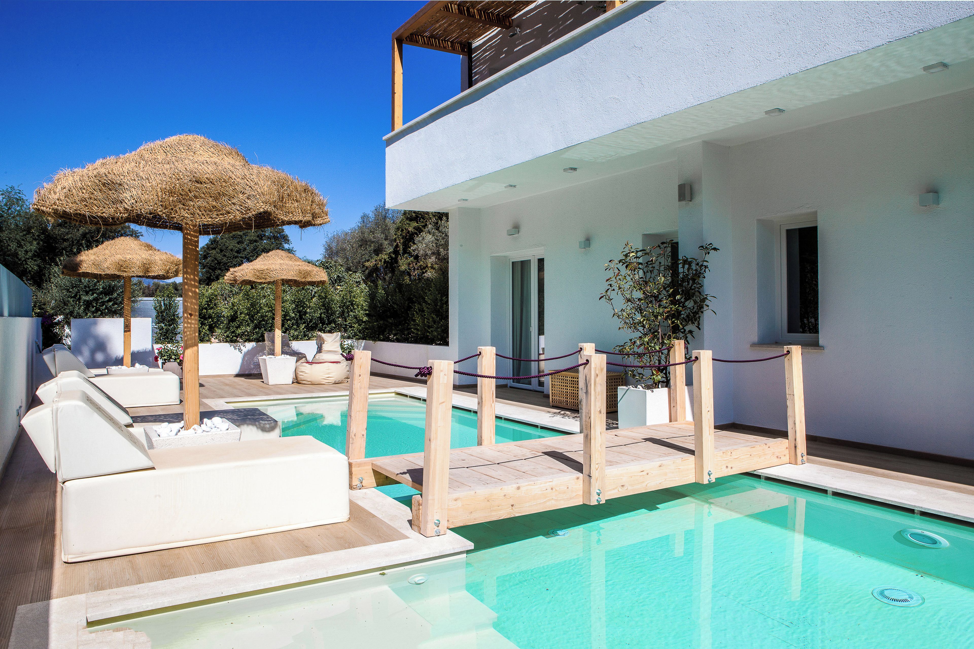 A pool area with lounge chairs and thatched umbrellas, wooden deck, and a white building with a balcony.