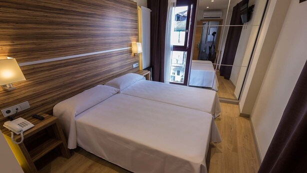 A hotel room with a wooden headboard, a bed with white linens, a bedside table with a lamp and a phone, and a view of the street through a window. - Hotel Novo Candido (Ourense)