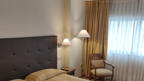 Una habitación de hotel con dos camas, una silla, una mesita de noche y un ventanal con cortinas.