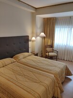 Una habitación de hotel con dos camas, una silla, una mesita de noche y un ventanal con cortinas.