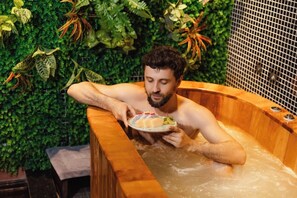 Een persoon in een houten jacuzzi, met een bord eten, omringd door weelderige groene begroeiing.
