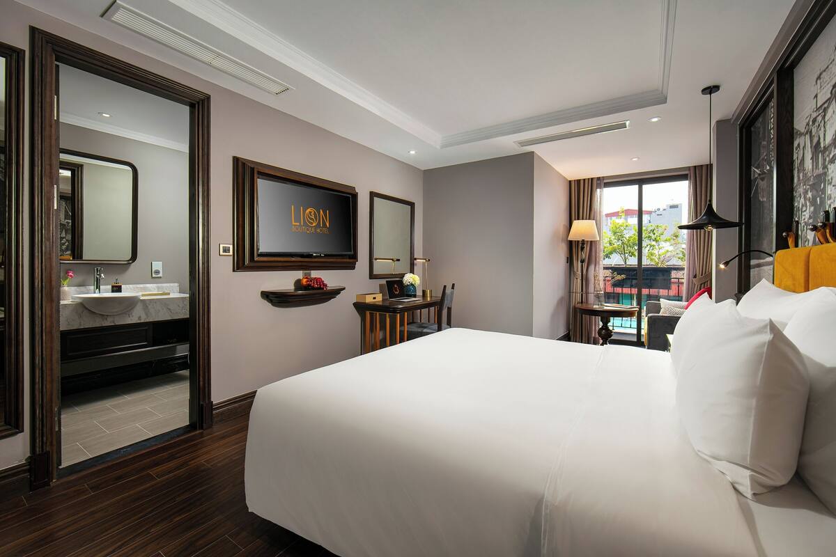 Una habitación de hotel con una cama, televisión, escritorio con lámpara, ventana con vistas y baño con lavamanos y espejo.