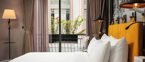 A hotel room with a bed, a lamp, a window with curtains, a view of the outside, and a wall with a picture and a shelf with books.
