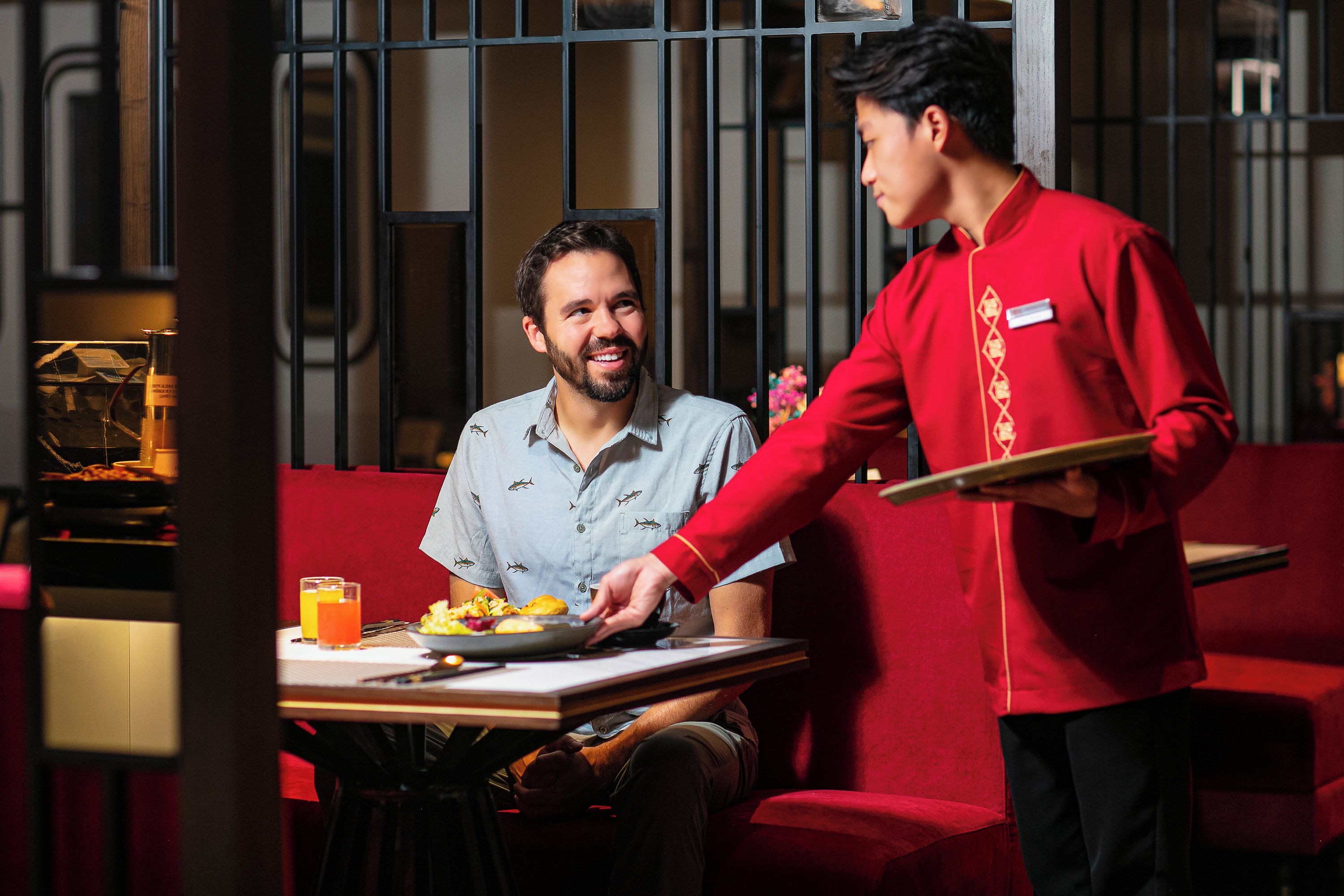 A dining area with a table set for two, a waiter in a red uniform, and a red cushioned booth in the background.