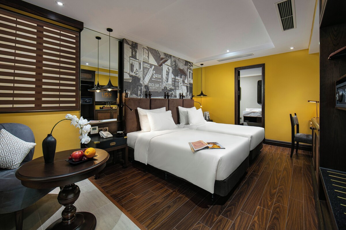 Ein Hotelzimmer mit einem großen Bett, einem Esstisch, einem Stuhl, einem kleinen Tisch mit einer Vase und Früchten sowie einem großen Spiegel.