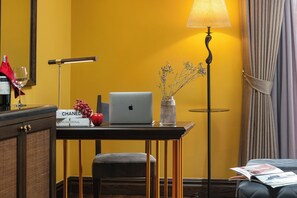 Une chambre d’hôtel avec un mur jaune, un bureau avec un ordinateur portable, une lampe, un vase contenant des fleurs séchées, une boîte Chanel, un verre à vin et un tissu rouge.