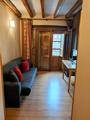 A room with a wooden floor, a sofa with red cushions, a wooden door, and a window with a view of buildings. - Hotel San Roque (Reinosa)
