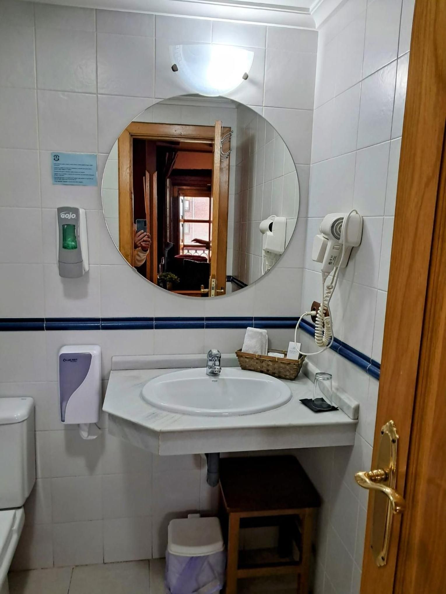 Un baño con un lavamanos blanco, un espejo, un secador de pelo, un secador de manos y un cubo de basura.