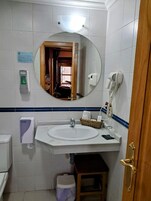 Un baño con un lavamanos blanco, un espejo, un secador de pelo, un secador de manos y un cubo de basura.