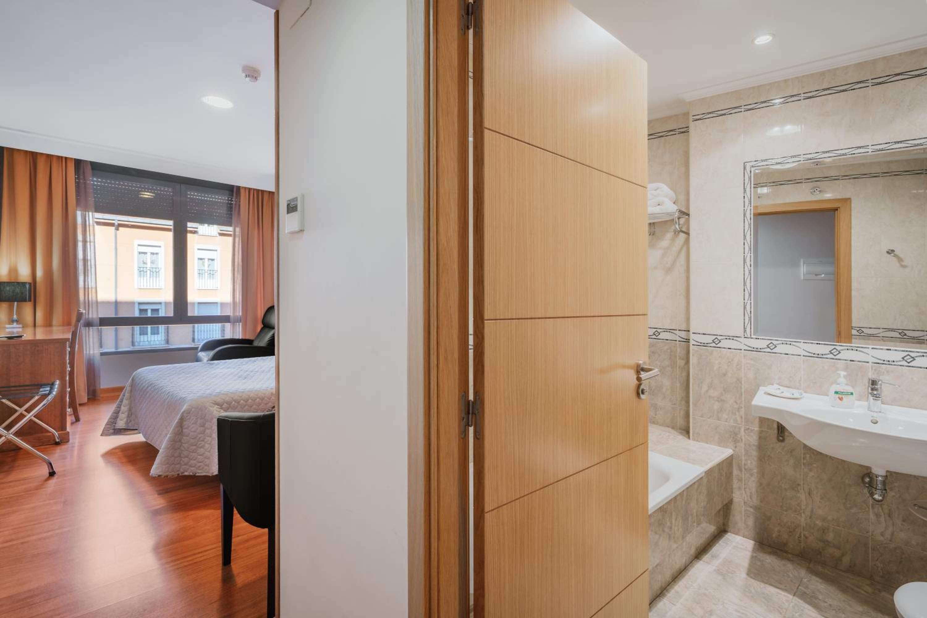 Habitación de hotel con cama, escritorio, silla, baño con lavamanos y espejo, y vistas al exterior.