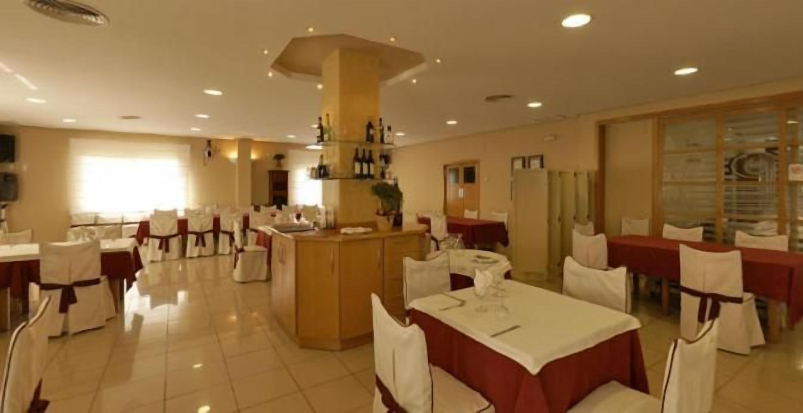 Un comedor amplio con múltiples mesas y sillas, una zona de bar central y un gran ventanal que proporciona luz natural.