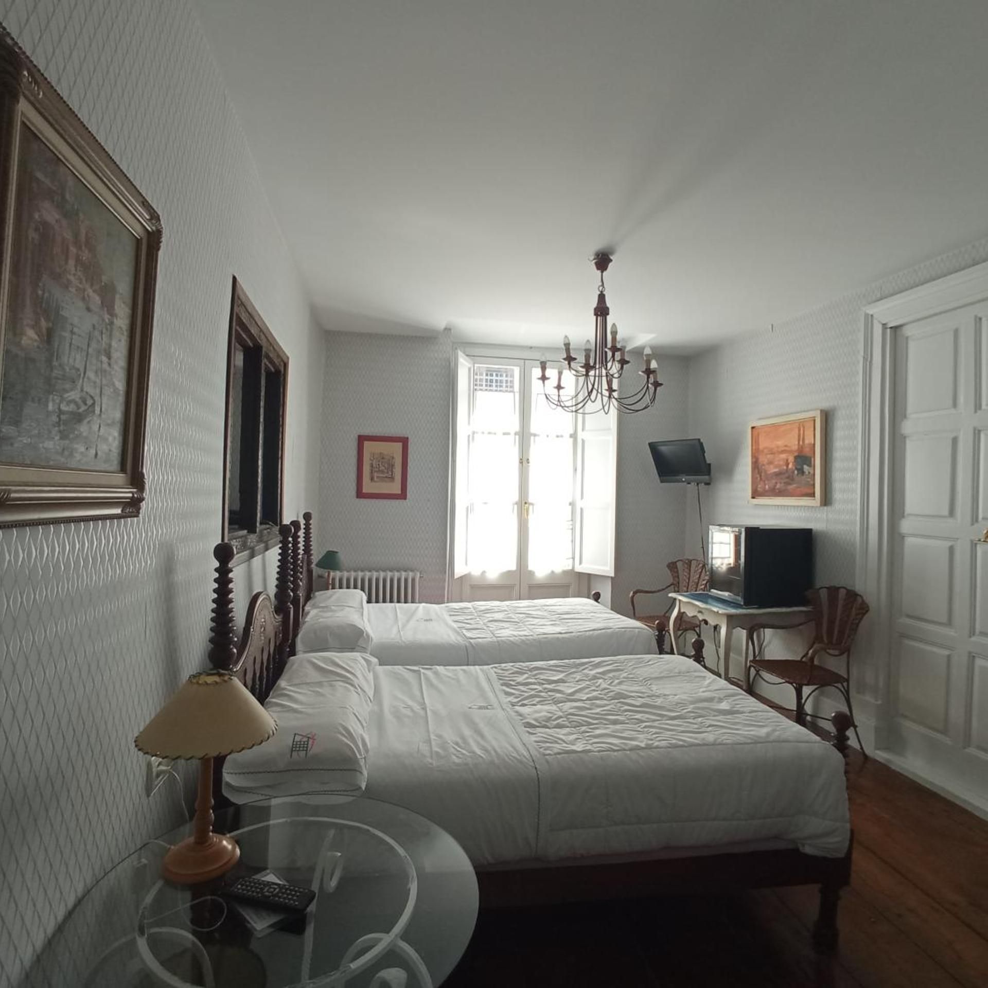 A hotel room with two beds, a chandelier, a television, and a window with natural light.