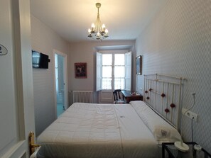 A hotel room with a bed, a desk, a chair, a radiator, a window, and a chandelier. - Hotel Ormazabal (Bergara)