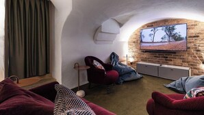 Un salon chaleureux avec un mur en pierre, un téléviseur à écran plat fixé au mur, un fauteuil rouge et un canapé agrémenté d’un plaid à motifs.