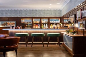 Een bar met houten lambrisering, groene barstoelen en diverse flessen op planken.