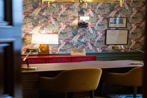 Een kamer met behang met patroon, een bureau met twee stoelen, een lamp en een rode kast.