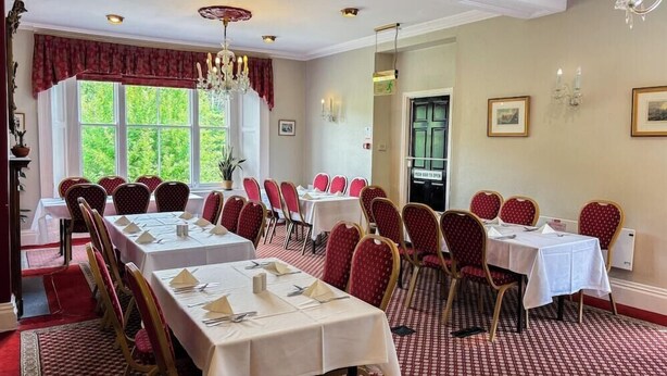 Uma sala de jantar formal com várias mesas e cadeiras, grandes janelas com cortinas e um lustre.