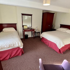 Ein Hotelzimmer mit zwei Betten, einem Schreibtisch mit Spiegel, einem Kleiderschrank und einem kleinen Tisch mit Lampe.